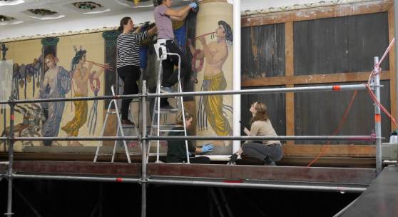 Vier Restauratorinnen nehmen ein großformatiges Gemälde für den Transport und die Bearbeitung von der Wand und drehen es vorsichtig auf eine große Rolle. Foto: Martina Schrei